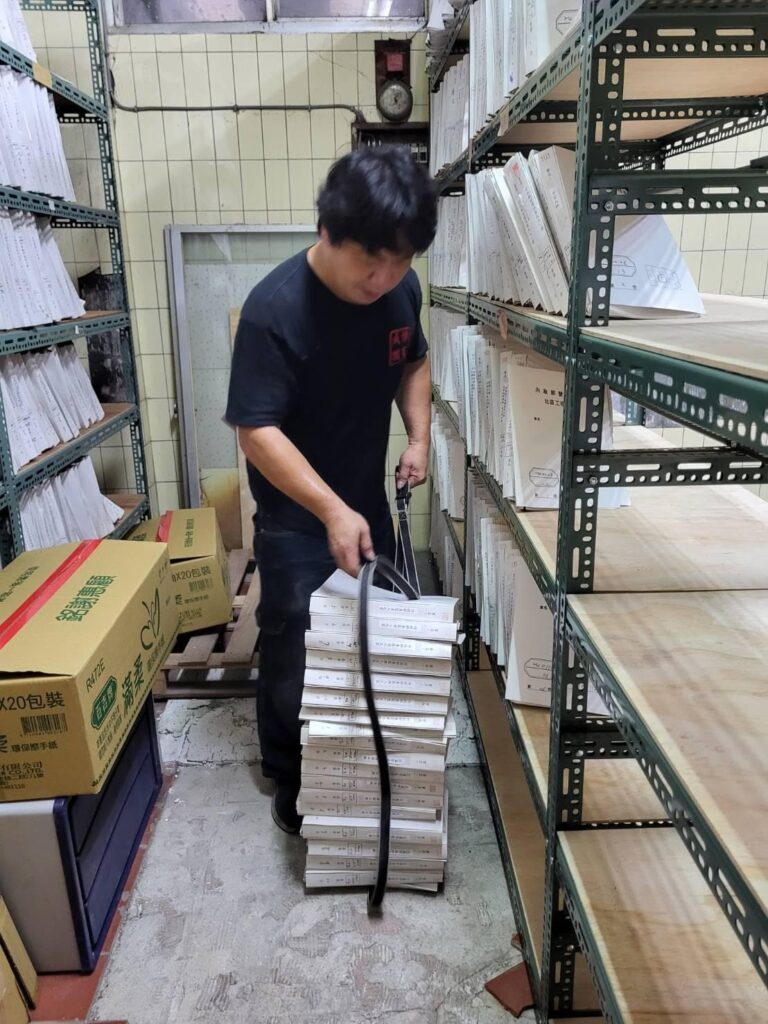 成傑搬家工作人員在倉庫整理一疊白色扁平包裝盒,貨架上擺滿各式文件紙品